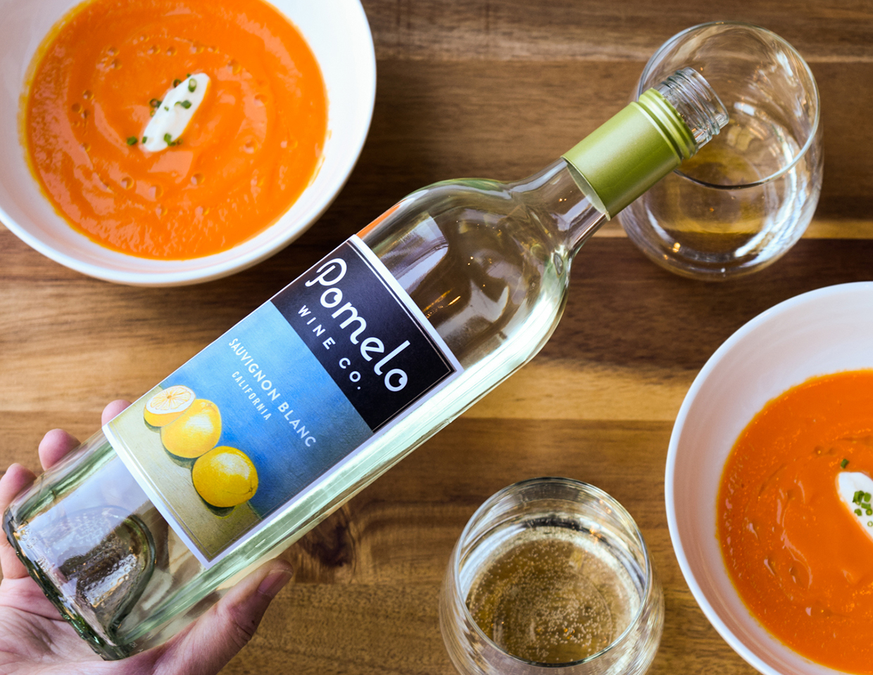 pouring a bottle of Pomelo sauvignon into a glass with bowls of tomato soup on a table